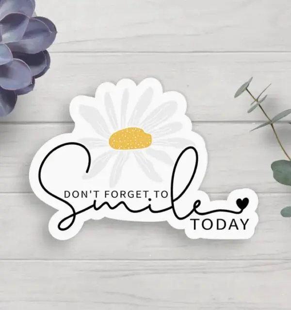 A sticker with the message "Don't forget to smile today" in cursive font, accompanied by an illustration of a daisy, on a light wooden background with leaves.