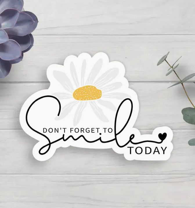 A sticker with the message "Don't forget to smile today" in cursive font, accompanied by an illustration of a daisy, on a light wooden background with leaves.