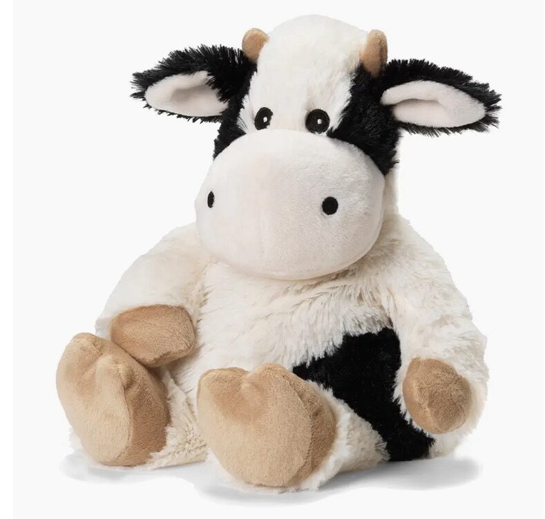 The Black And White Cow Warmie is a plush toy with black and white fur, tan hooves, and large round eyes, sitting upright against a white background.