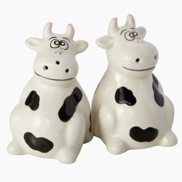 Two ceramic figures from the Cows Salt And Pepper Shakers set, featuring black and white cartoonish cows, are sitting side by side against a plain background.