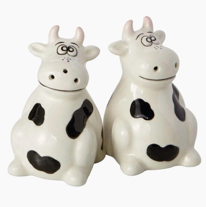 Two ceramic figures from the Cows Salt And Pepper Shakers set, featuring black and white cartoonish cows, are sitting side by side against a plain background.