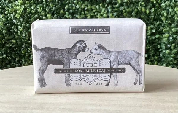 A bar of Beekman 1802 Fragrance Free Goat Milk Bar Soap 9oz rests on a wooden surface, showcasing an illustrated label with two goats and highlighting its sulfate and paraben-free qualities.
