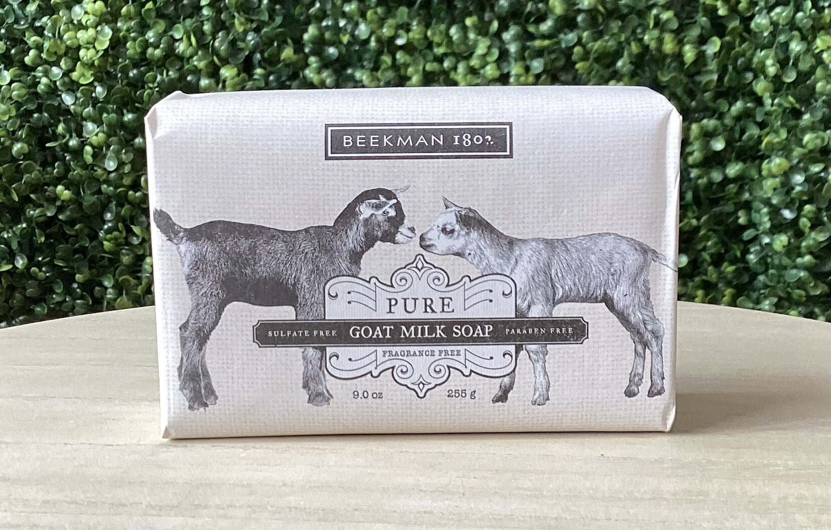 A bar of Beekman 1802 Fragrance Free Goat Milk Bar Soap 9oz rests on a wooden surface, showcasing an illustrated label with two goats and highlighting its sulfate and paraben-free qualities.