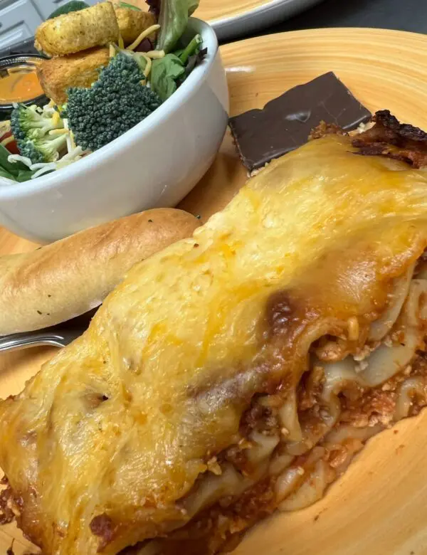 A serving of the Lasagna Dinner on a wooden plate, complete with a side salad, breadstick, and a piece of chocolate.