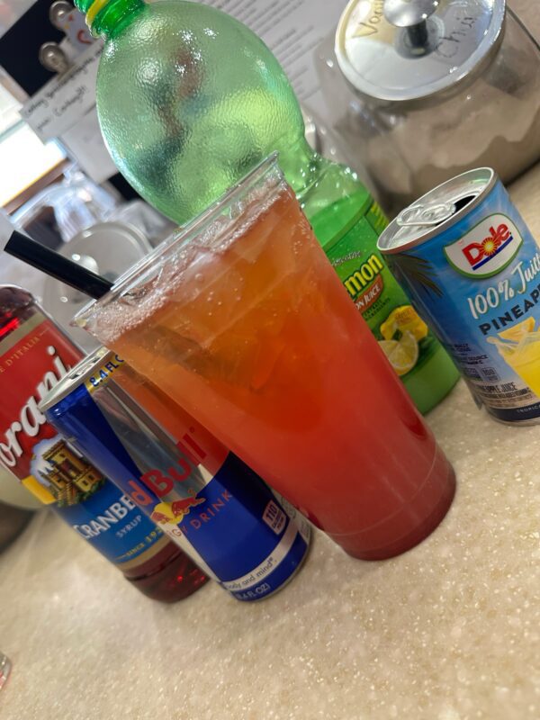 A Grinchberry Refresher (Red Bull) in a clear cup with a straw, surrounded by bottles and cans of various beverages, on a countertop.