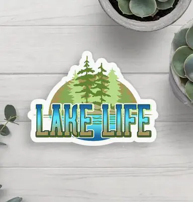 Sticker with "Lake Life" text, featuring illustrations of trees and a lake, placed on a light wooden surface, surrounded by potted succulent plants.