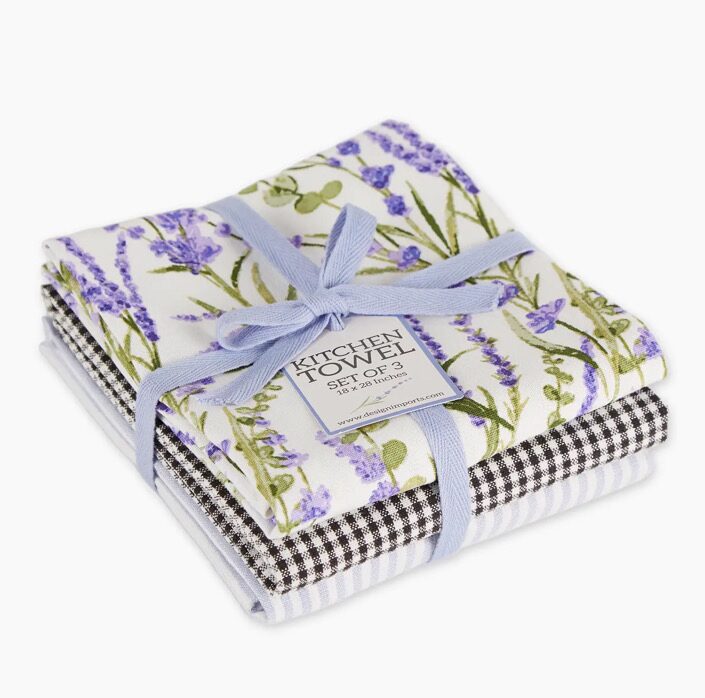 The Lavender Fields Set Of 3 includes kitchen towels elegantly folded and tied with a light blue ribbon. The top towel showcases a lavender floral design, while the remaining two feature black and white checkered and striped patterns.