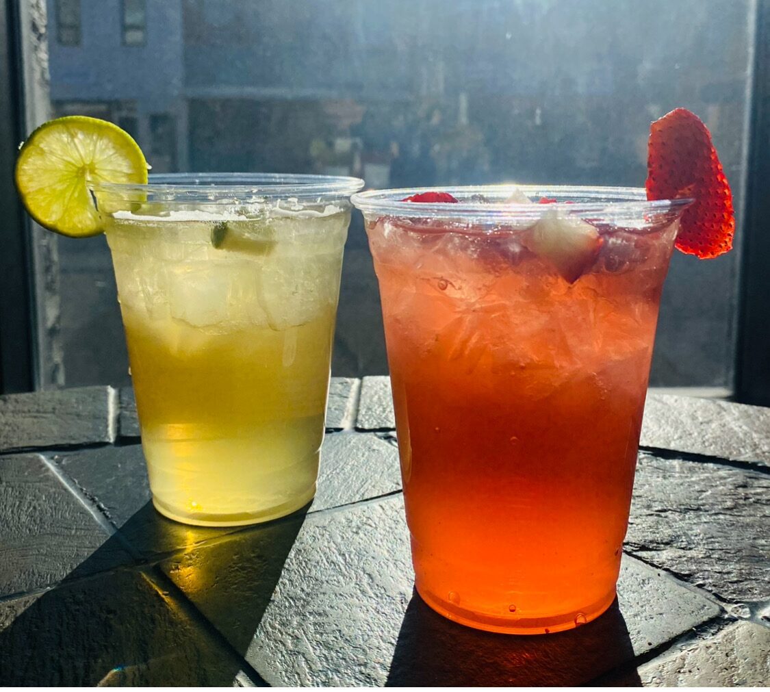 Two drinks in plastic cups on a table. The left drink is yellow with a lime slice, and the right is red with a strawberry garnish. Sunlight casts shadows on the table.