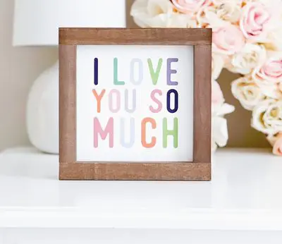 A piece of framed wall decor placed on a table reads "I LOVE YOU SO MUCH" in vibrant letters.