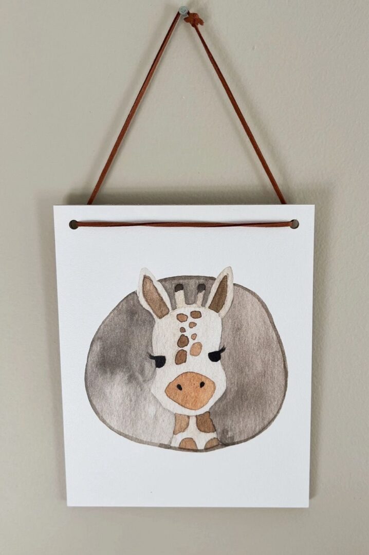 The Giraffe Circle Hanging Wall Art, featuring a watercolor painting of a giraffe's head with closed eyes on a white canvas, is elegantly suspended by a brown string against a light gray wall. Perfect for nursery decor.