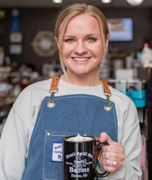 Person wearing a denim apron holds a mug with latte art, standing inside a coffee shop.